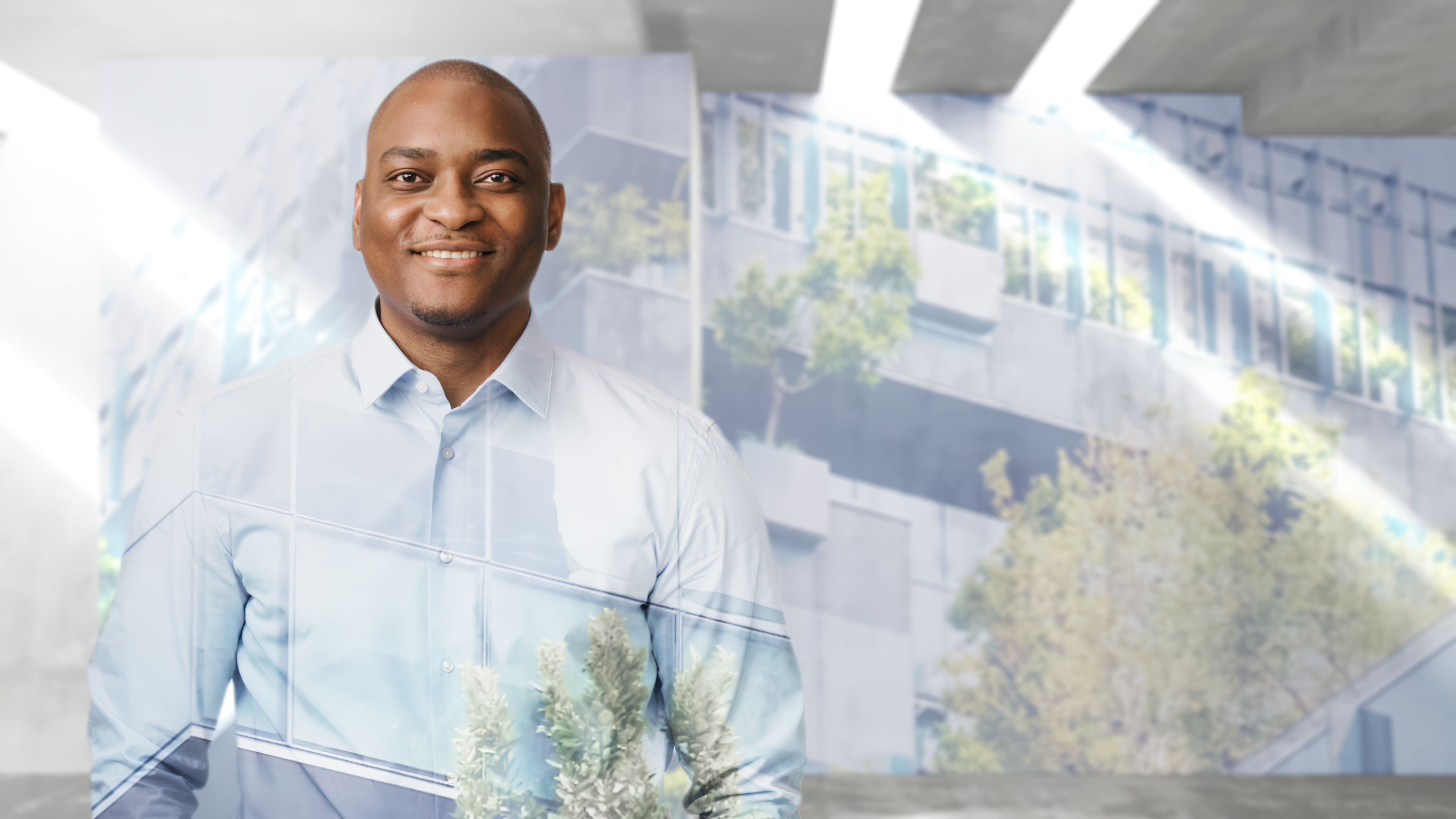 A friendly, smiling man in a light blue shirt stands in front of the soft-focus background of a modern, light-filled office building with trees.