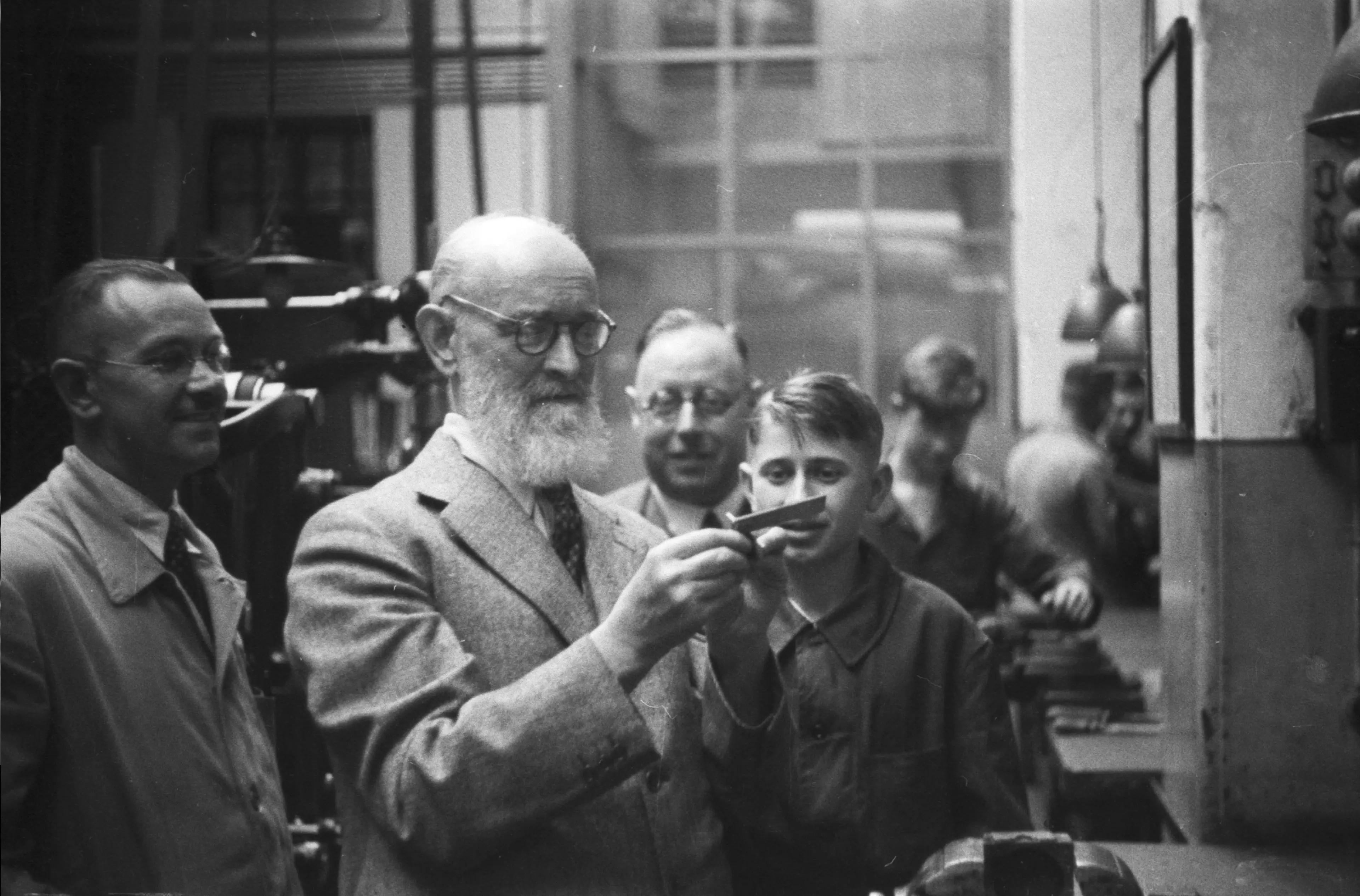 Historische Schwarz-Weiß-Aufnahme von Robert Bosch mit Bart und Brille in einer Werkstatt. Er ist von Mitarbeitern umgeben und begutachtet prüfend ein kleines Werkstück.