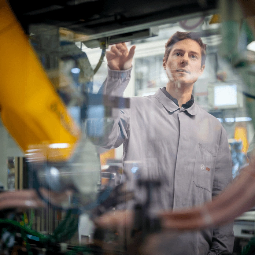 Ein Bosch-Mitarbeiter in einem grauen Arbeitskittel steht in einer Fertigungsanlage und blickt auf eine Maschine. Die Aufnahme ist durch eine Glasscheibe gemacht, in der sich ein gelber Roboterarm spiegelt.