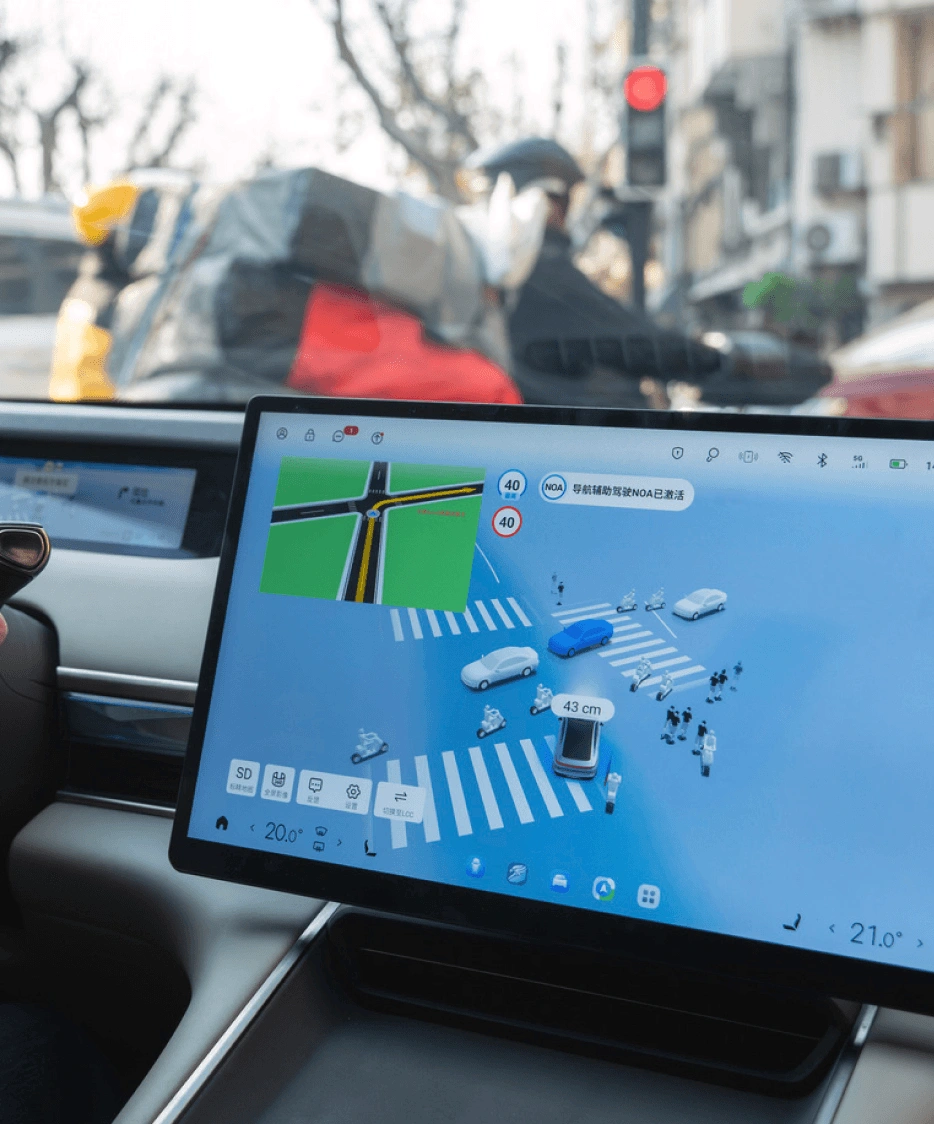 Nahaufnahme des Armaturenbretts eines Autos. Die Hände der Fahrerin sind am Lenkrad. Ein großer Touchscreen in der Mitte zeigt das ADAS-System, eine 3D-Visualisierung der Fahrzeugumgebung, einschließlich anderer Verkehrsteilnehmer und der Fahrspurmarkierungen.