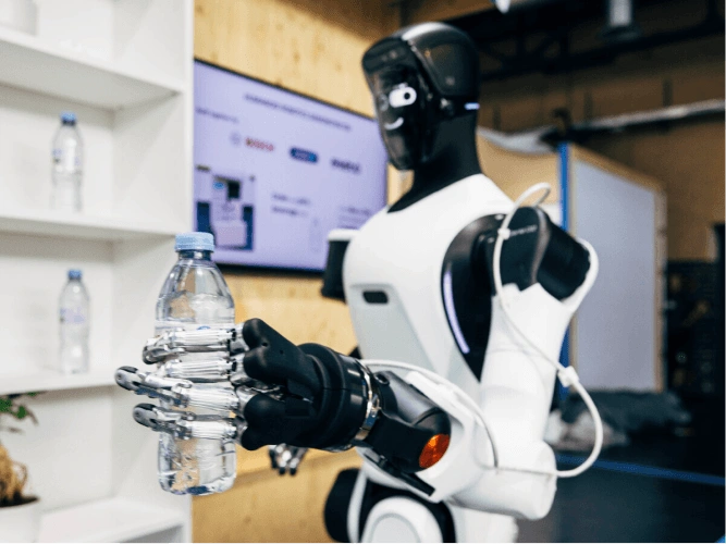 Ein humanoider Roboter mit weiß-schwarzem Körper hält mit seiner metallischen Roboterhand eine Plastikwasserflasche. Im Hintergrund sind weiße Regale zu sehen.